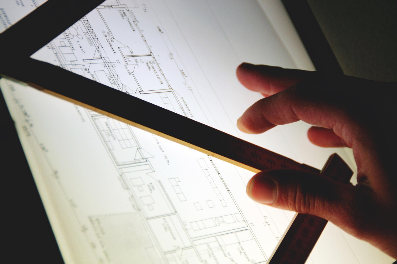 Une main tient une règle posée sur un plan de construction éclairé, pour relever précisément les postes d'un devis de travaux