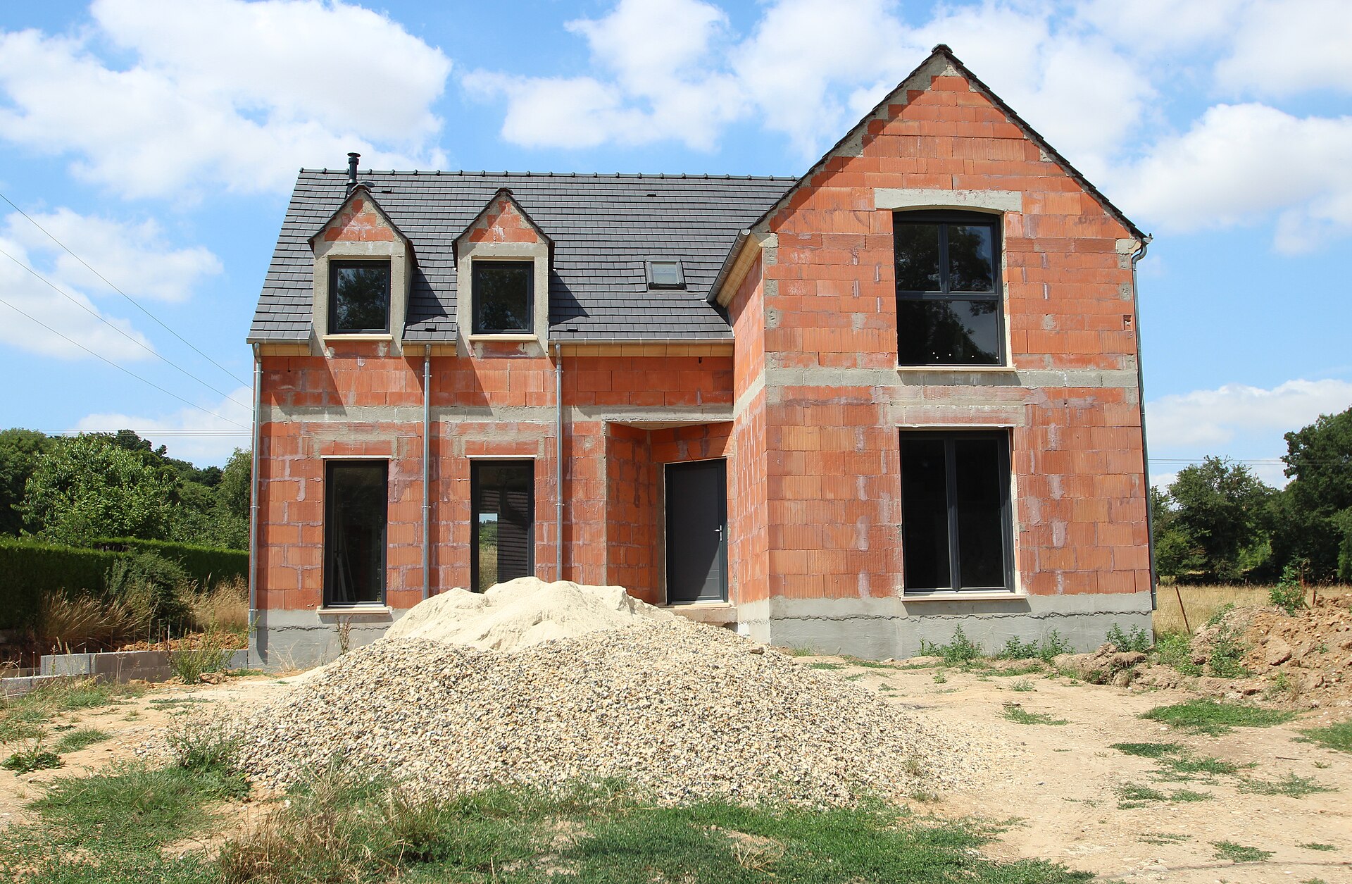 maison neuve en briques avec une toiture en ardoise percée de trois lucarnes, terrain encore en chantier avec un tas de gravier